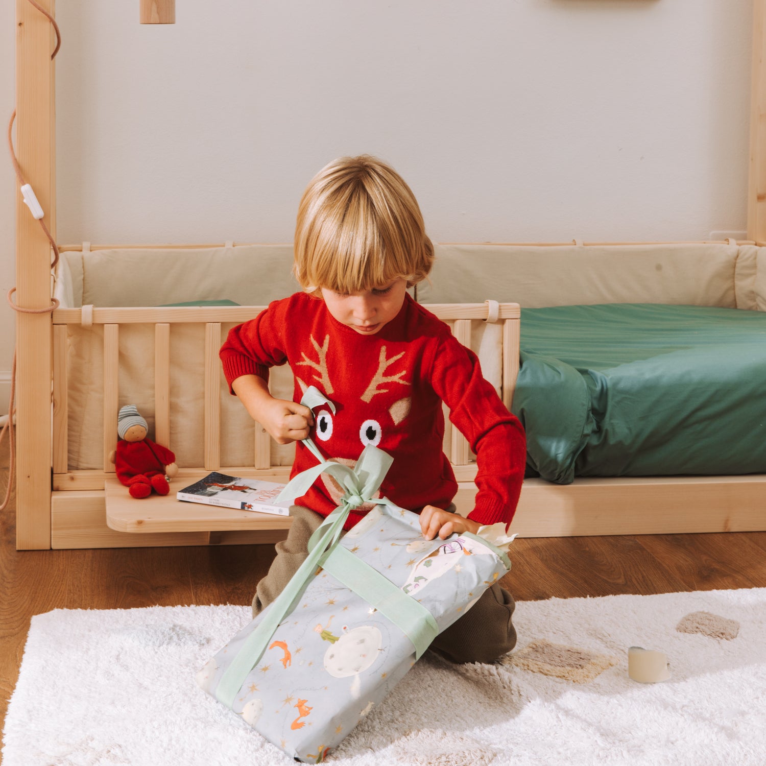 Un bambino con un pullover rosso apre un regalo. Sullo sfondo un letto Montessori nabè con paracolpi, barriere e comodino. 